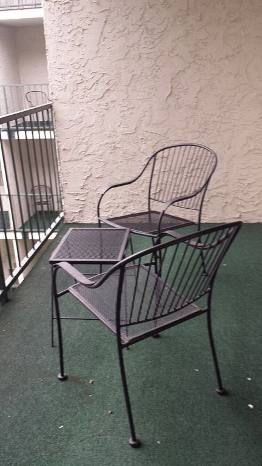 Conjunto de mesa e cadeira de metal numa varanda com parede texturizada e guarda-corpo de metal.