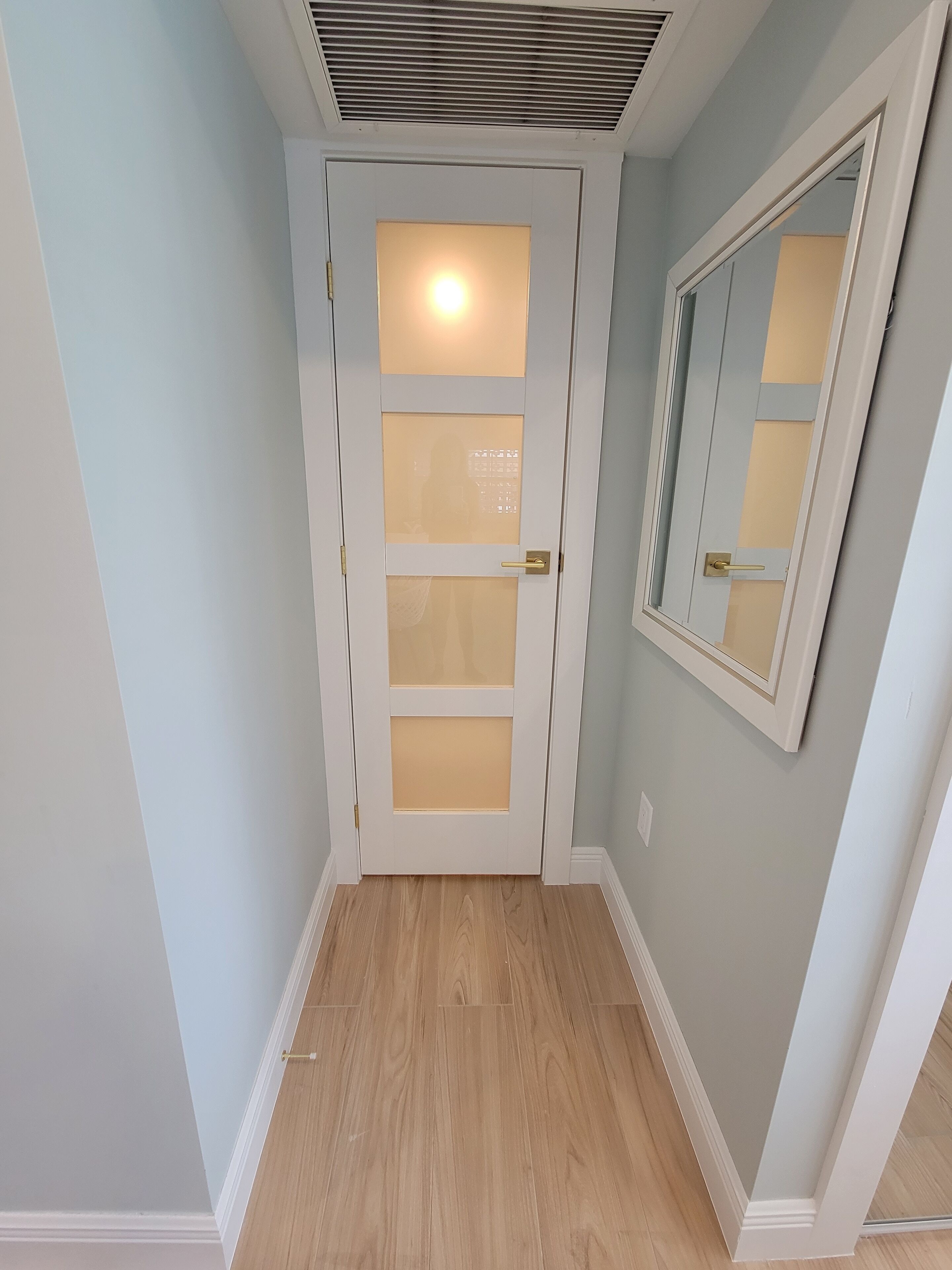Un pasillo con una puerta blanca que tiene paneles de vidrio esmerilado, un interruptor de luz y una rejilla de ventilación arriba.