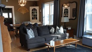 Ein Wohnzimmer mit einem dunkelgrauen Sofa, einem Holztisch und einem gemusterten Sessel. Eine Pendelleuchte hängt von der Decke, und im Hintergrund ist ein Essbereich zu sehen.