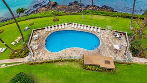 Una piscina con cubierta de piedra y sillas de descanso, rodeada de césped cuidado y palmeras, con vista al océano.