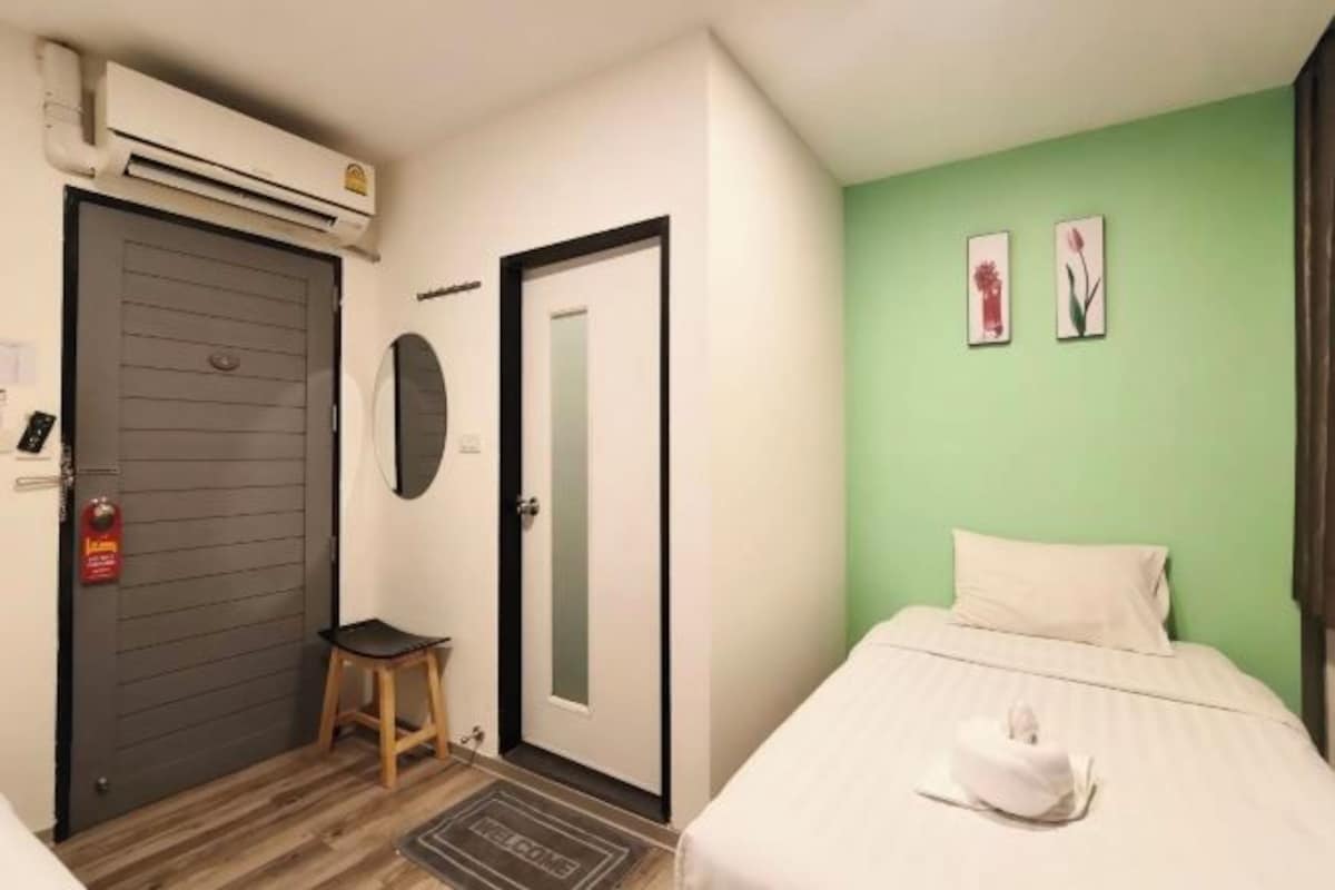 A hotel room with a bed, a door, a small stool, and a wall-mounted air conditioning unit.