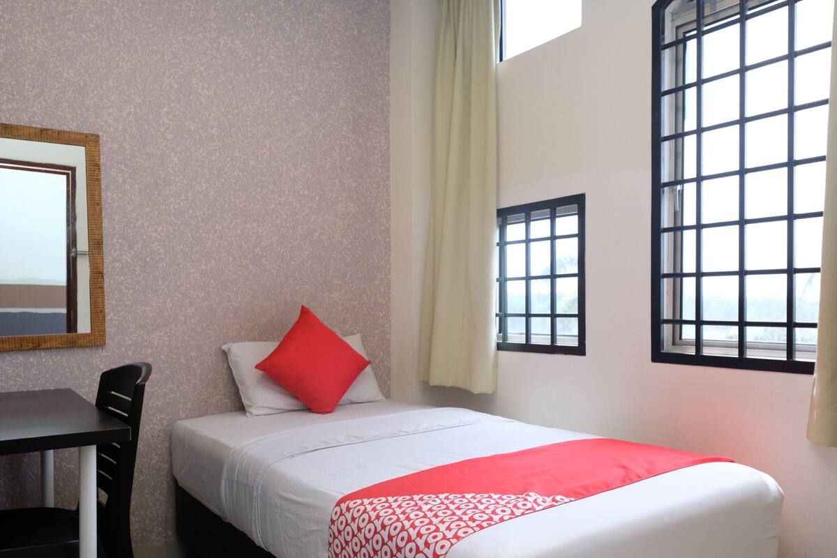 Una cama individual con un cojín rojo, una mesita y una silla en una habitación con ventana y espejo en la pared.