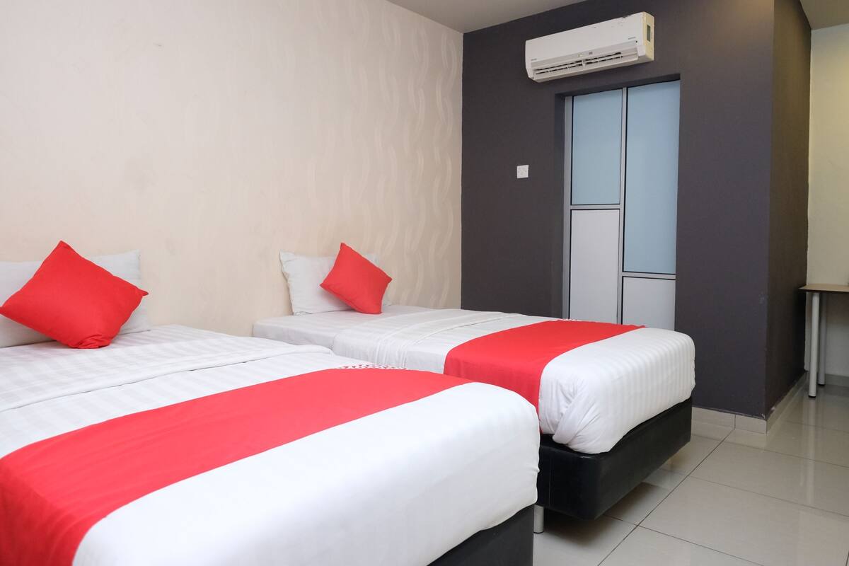 Habitación de hotel con dos camas, cada una con ropa blanca y detalles rojos, una pared gris y una ventana con vistas.