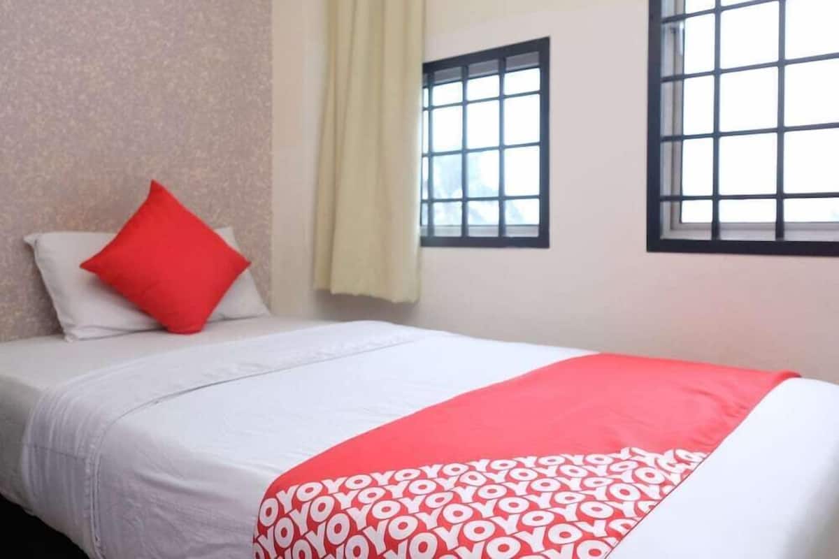 Una cama individual con una colcha blanca y roja de estampado, una almohada roja y una ventana con diseño de cuadrícula.