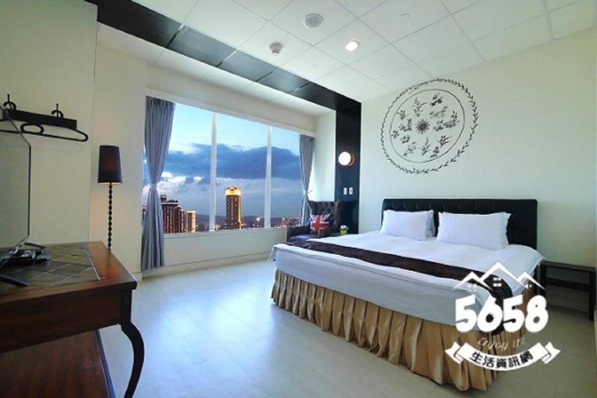 A hotel room with a large bed, a desk with a lamp, a view of the cityscape through the window, and a decorative wall piece.
