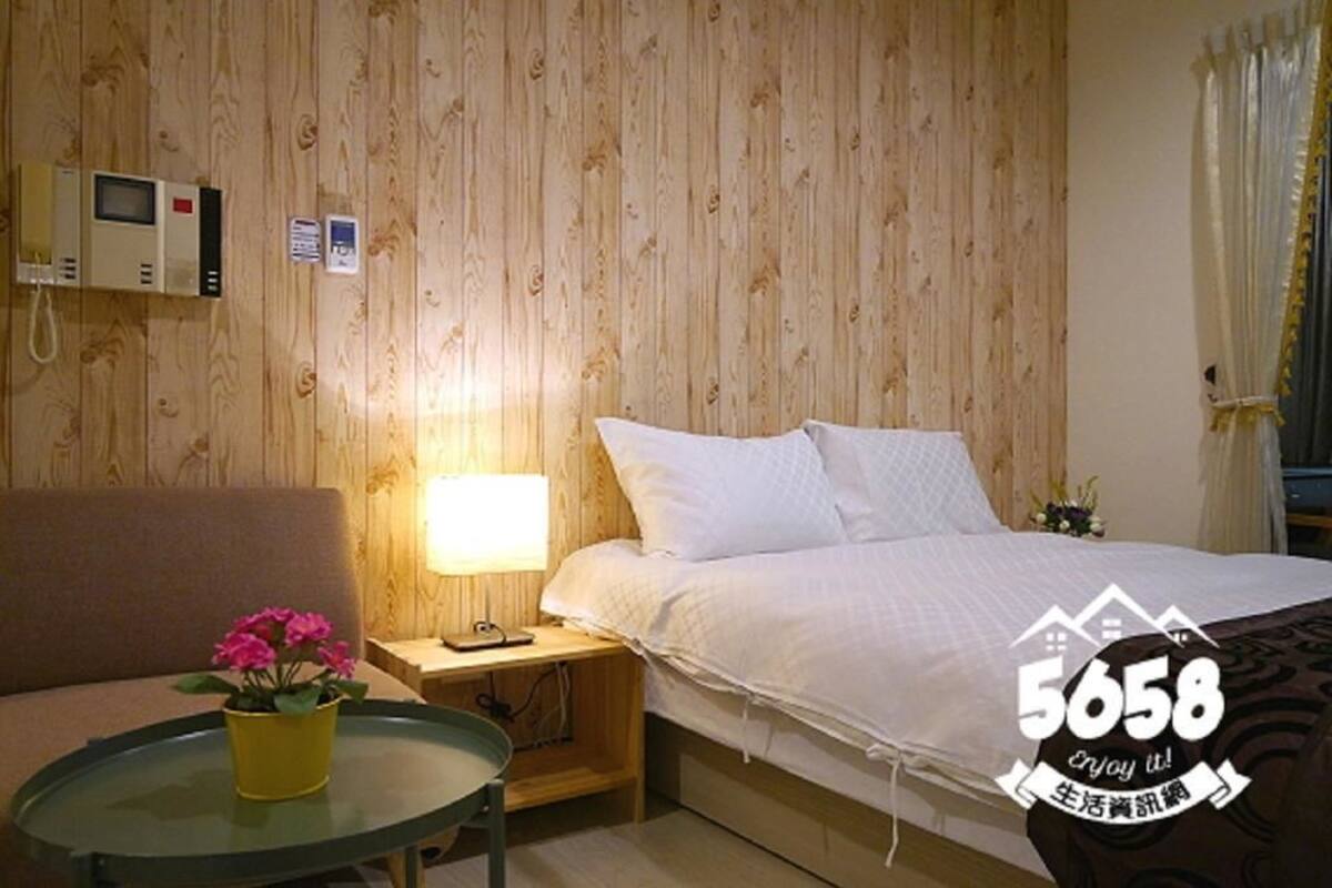A hotel room with a wooden panel wall, a bed with white bedding, a bedside table with a lamp, a small table with a yellow pot of flowers, and a TV on the wall.