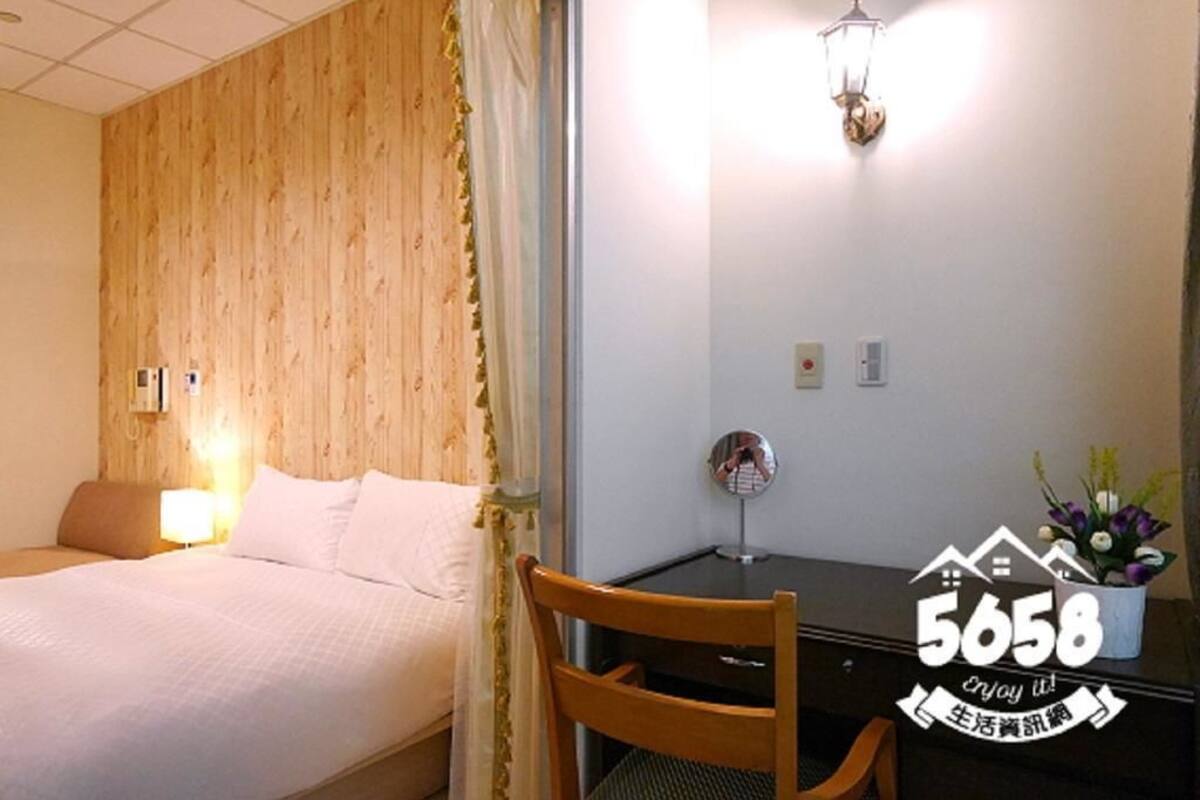 A hotel room with a wooden panel wall, a bed with white bedding, a desk with a chair, a wall-mounted lamp, and a small potted plant.