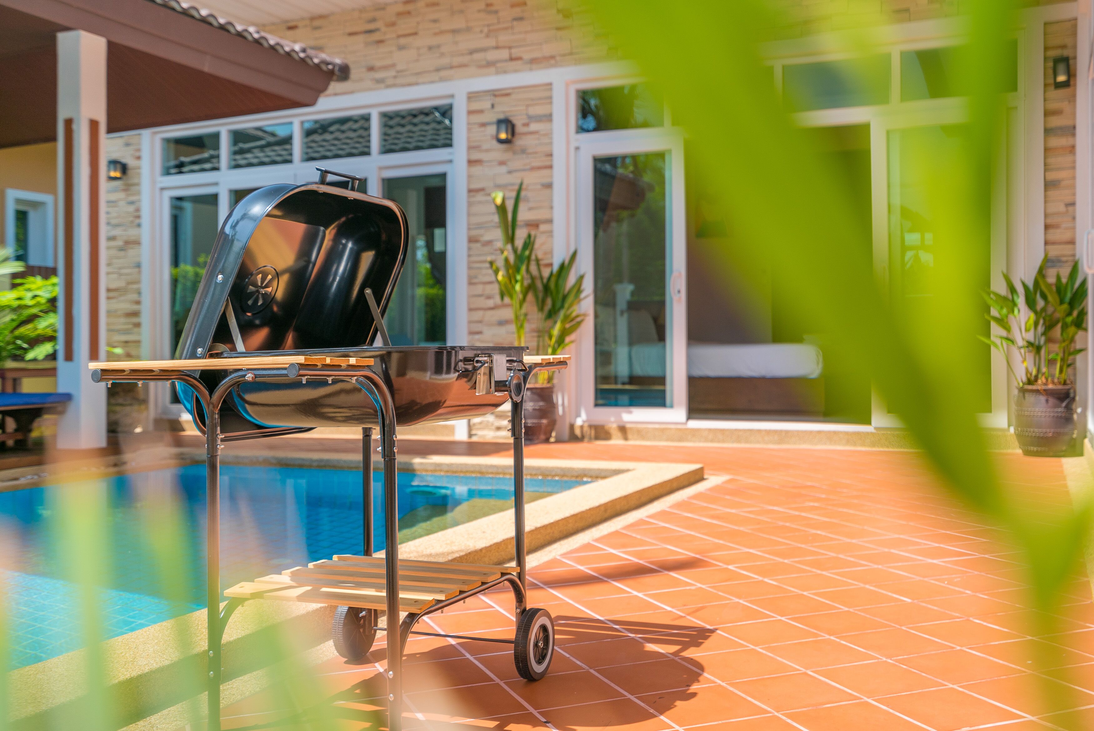Zona de piscina con un carrito para parrilla cerca, un patio con baldosas y un edificio con ventanas amplias.