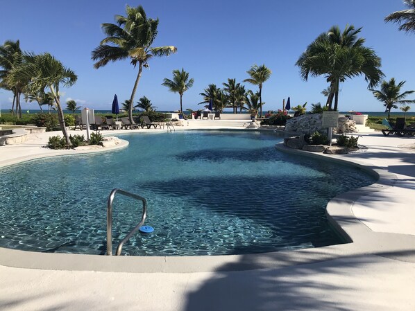 Relax by the pool surrounded by palm trees and lounge chairs, with the ocean visible in the background.