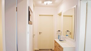Un pasillo estrecho con una puerta blanca, un aparador de madera con cajones, un espejo y un mueble pequeño con un lavabo.
