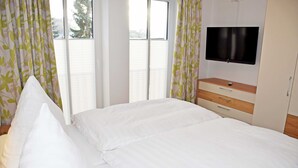 Un dormitorio con una cama, un televisor instalado en la pared y una ventana con cortinas.