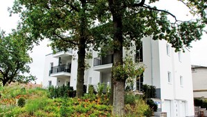 Un edificio moderno de dos pisos con balcones, rodeado de abundante vegetación y un jardín ajardinado.