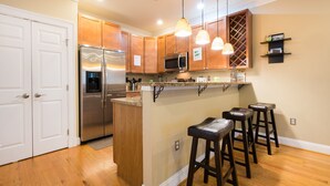 Une cuisine avec des meubles en bois, des appareils en acier inoxydable, un îlot central avec des tabourets, un éclairage suspendu et un meuble à vin.
