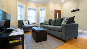 Un salon moderne avec un canapé gris, un pouf noir et un téléviseur à écran plat posé sur une table basse noire.