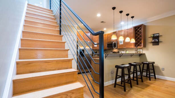 Un escalier en bois avec une rampe en métal mène à un espace cuisine doté de meubles en bois, d’un tabouret de bar et d’un éclairage suspendu.