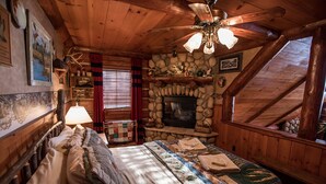 A cozy cabin-style bedroom with a stone fireplace, a bed with quilts, a wooden ceiling, and a window with curtains.