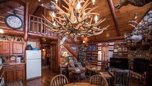 A rustic cabin interior with a wooden dining table, a kitchen area, a staircase, and a fireplace.