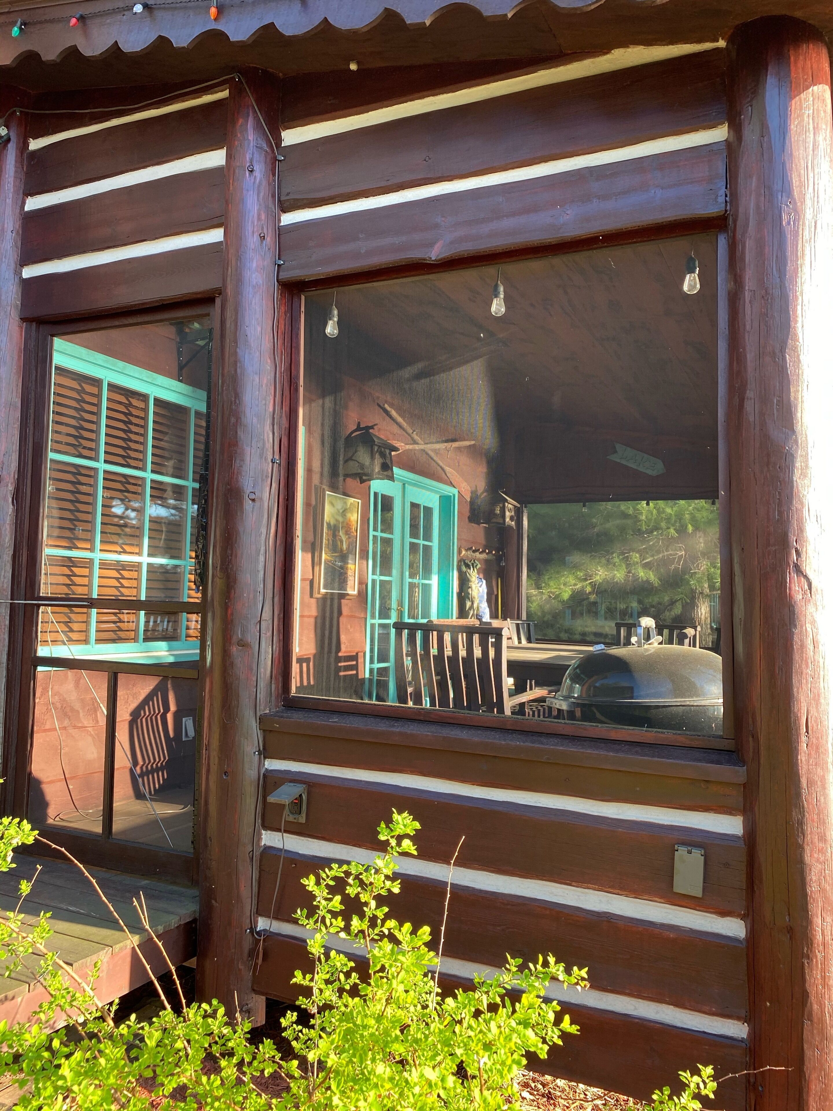 A rustic wooden cabin with a porch, a covered outdoor seating area, and a view of a turquoise building in the background.