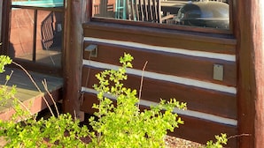 A rustic wooden cabin with a porch, a covered outdoor seating area, and a view of a turquoise building in the background.