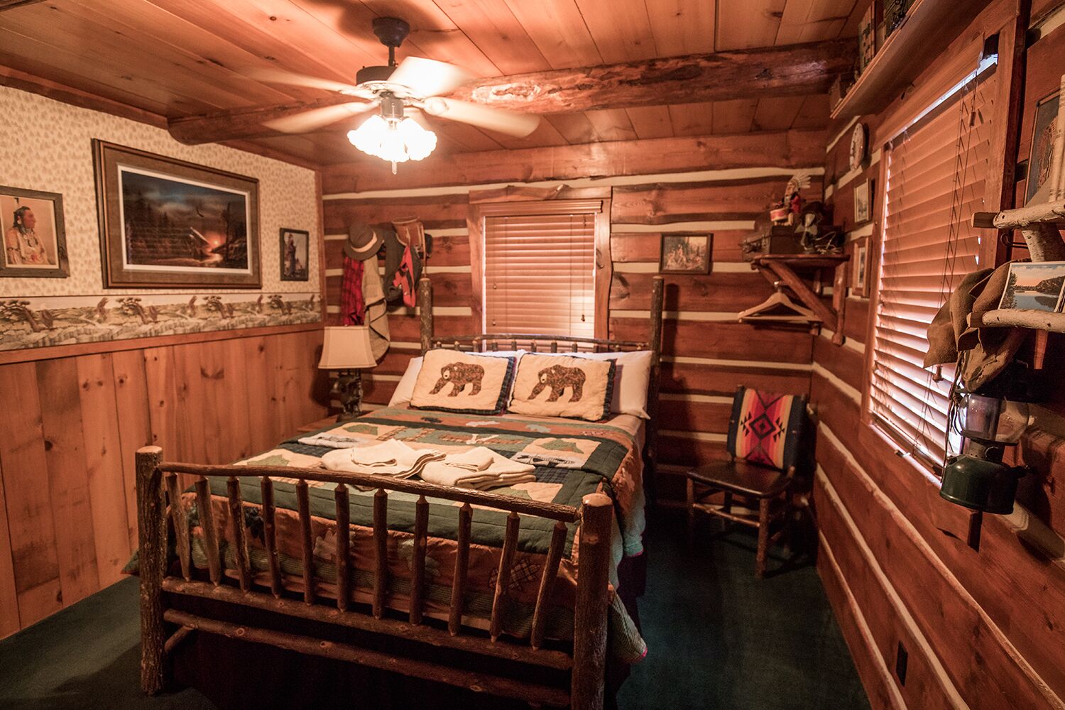 Een knus houten blokhutkamer met een houten bed, een plafondventilator en een rustieke inrichting, waaronder ingelijste foto's en een geborduurde sprei.