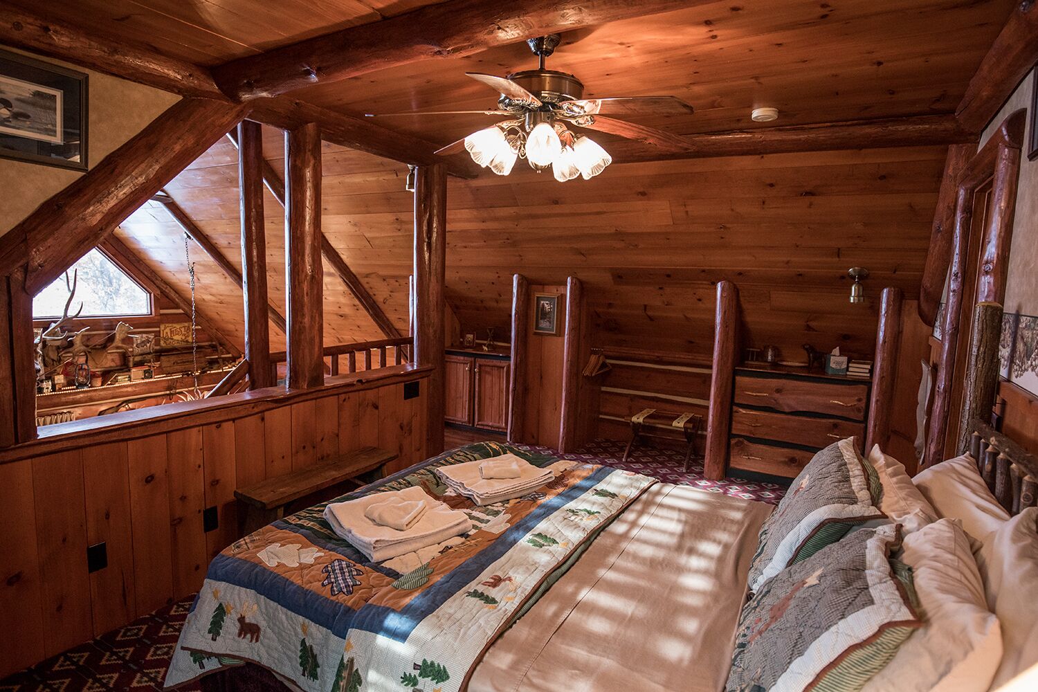 A cozy attic bedroom with a large bed, wooden beams, a ceiling fan, and a window overlooking a rustic cabin.