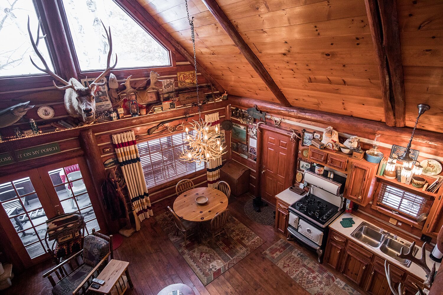 Interior rústico de una cabaña con comedor, cocina y un ventanal decorado con una cabeza de venado.