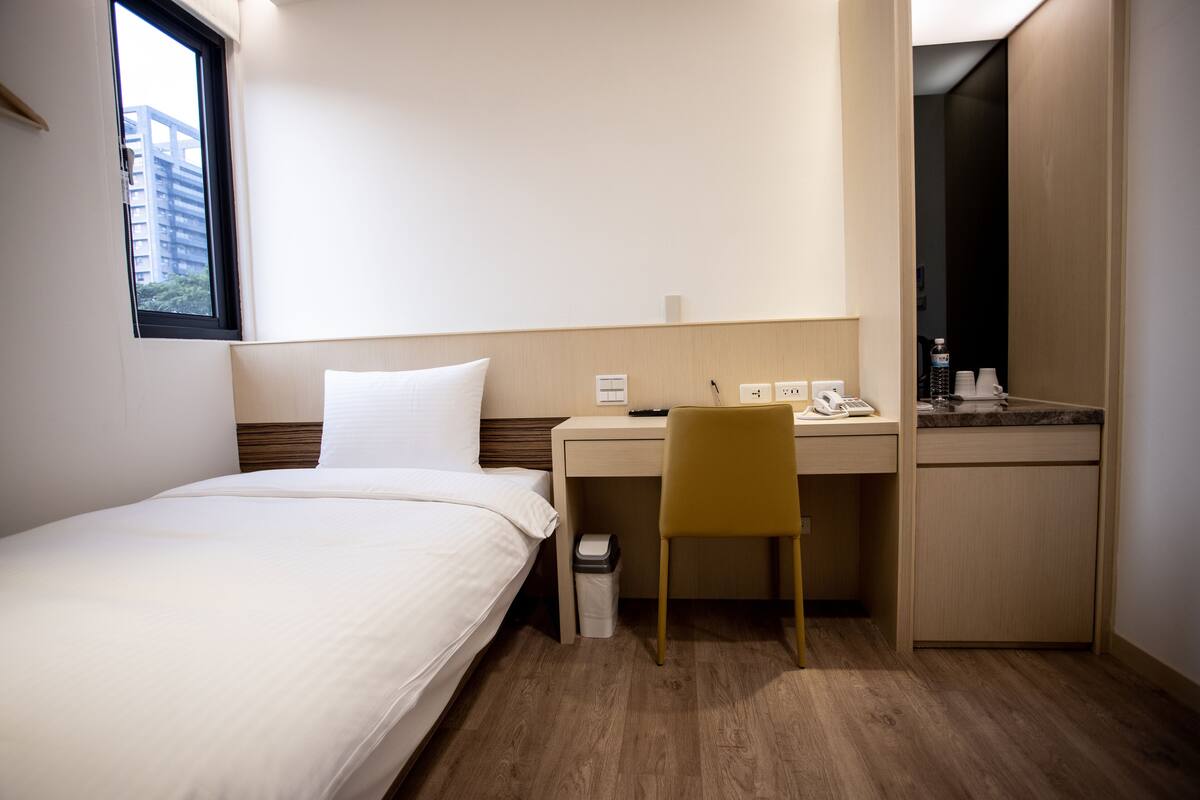 A hotel room with a bed, a desk, a chair, a small trash bin, and a kitchenette area.
