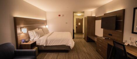 A hotel room with a bed, bedside table, lamp, TV, desk, chair, and a wooden floor.