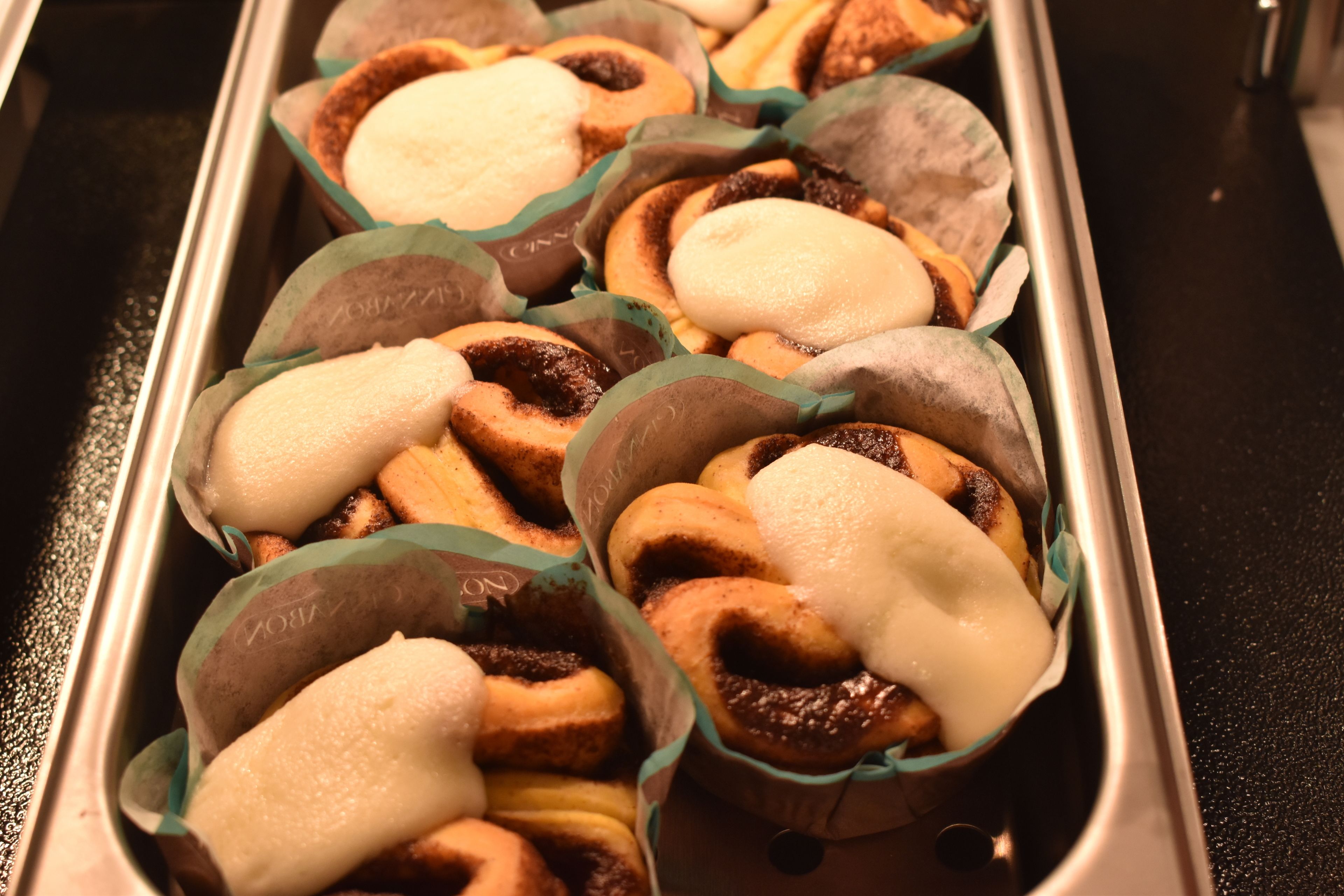 A tray of pastries with a variety of toppings, including white and chocolate glazes, arranged in a metal container.