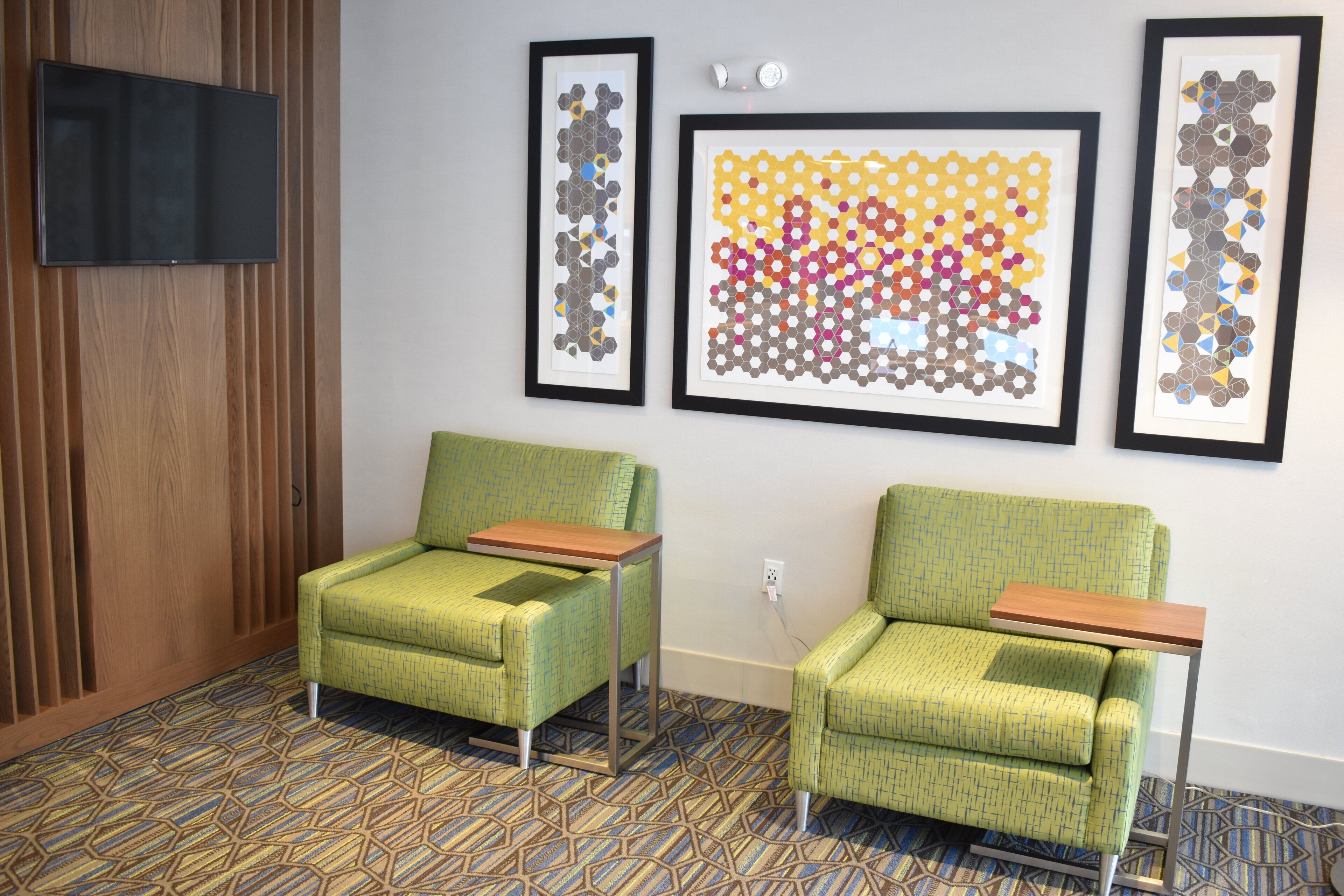 A room with two green armchairs, a flat-screen TV mounted on a wooden panel, and three framed geometric art pieces on the wall.