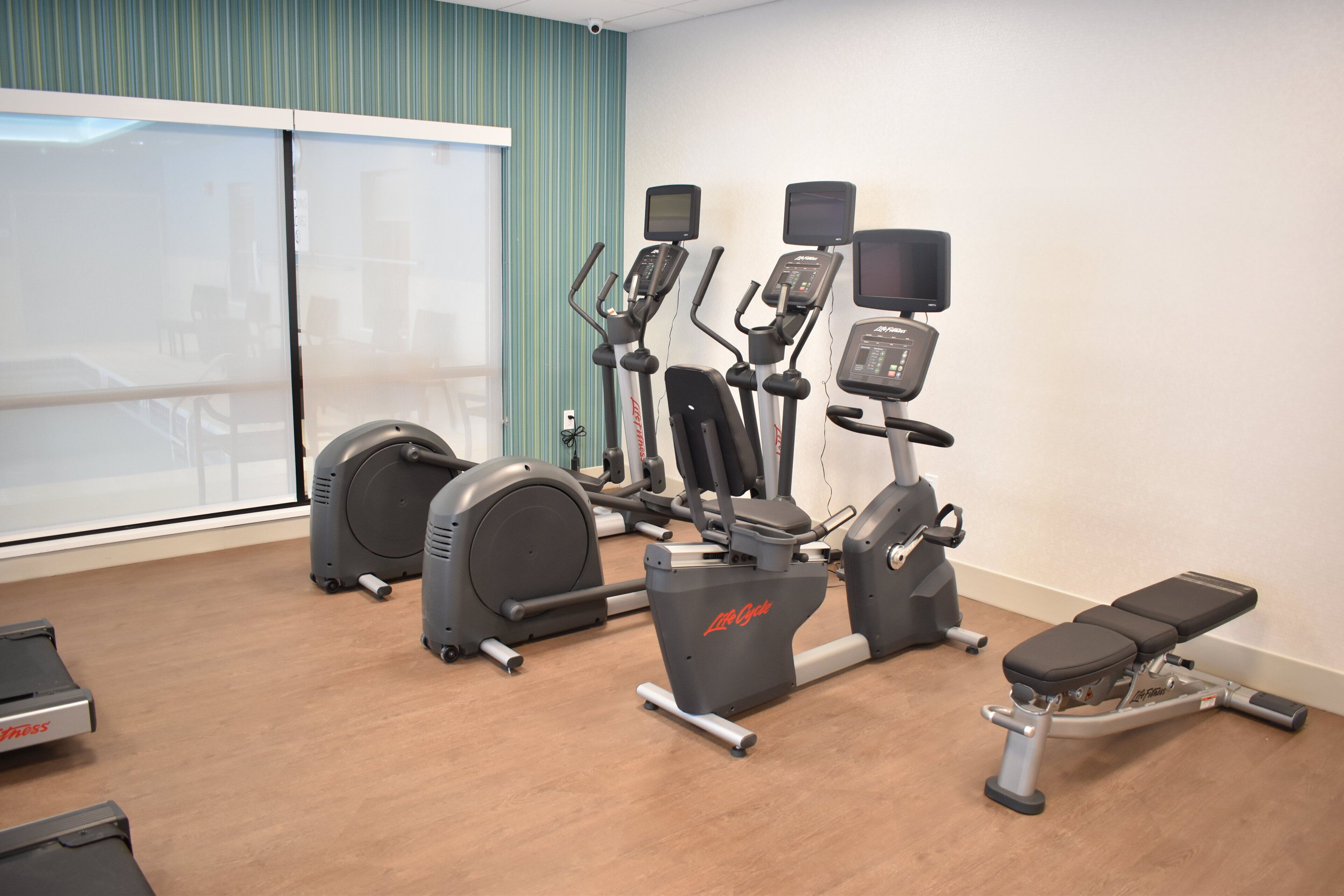 A gym with cardio machines, a bench, and a large window with curtains.