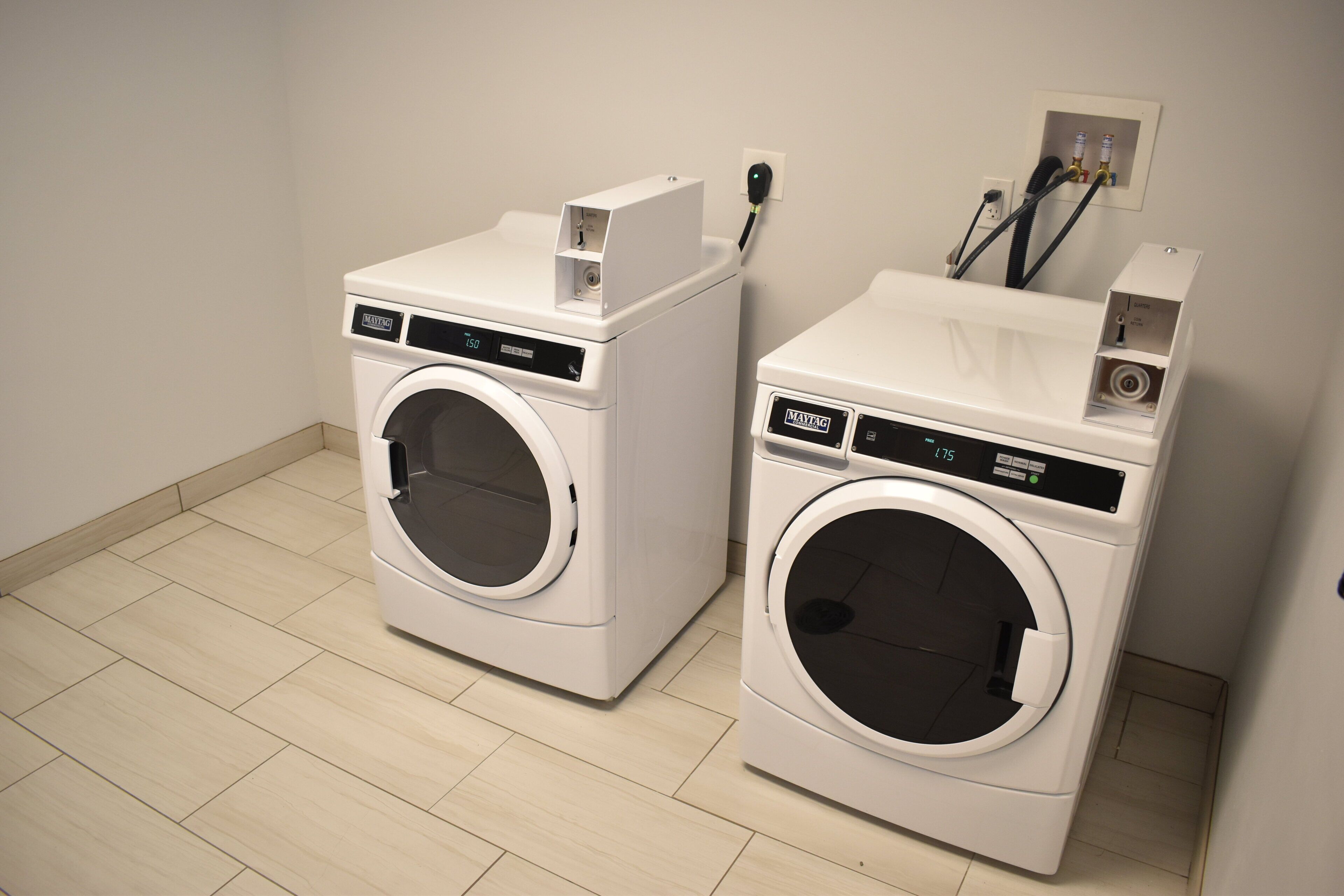 Two front-loading washing machines in a room with a tiled floor and a wall-mounted electrical outlet.