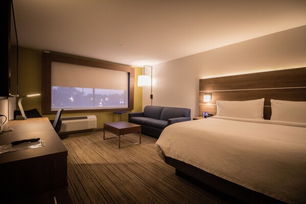 A hotel room with a bed, a sofa, a desk with a lamp, a TV, and a window with a view of the city. - Holiday Inn Express & Suites Boston South - Randolph by IHG (Randolph)