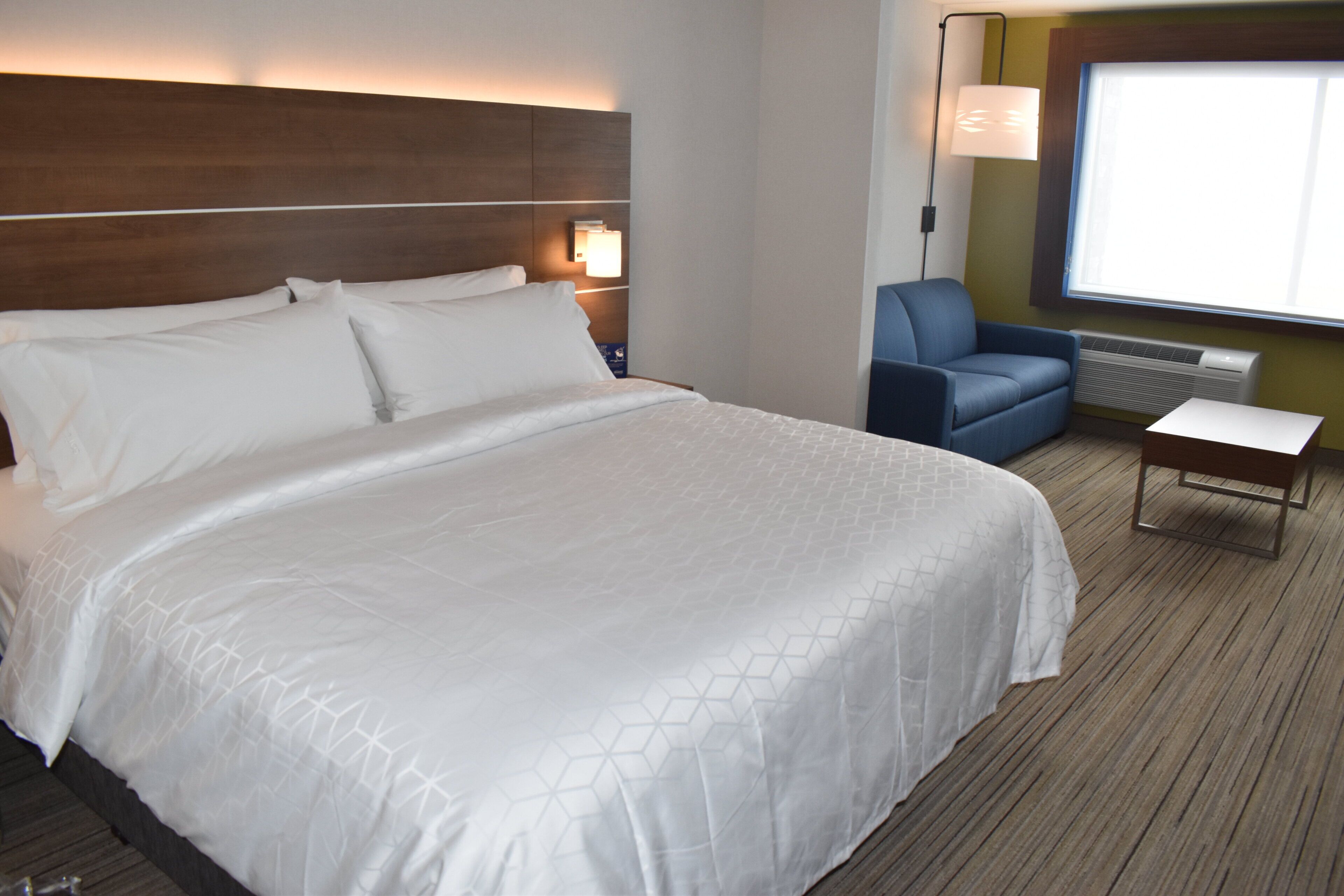 A hotel room with a large bed, a desk, a chair, a lamp, and a large window.