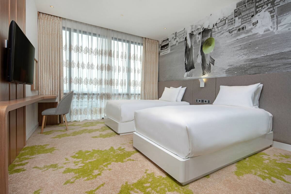 Une chambre d’hôtel avec deux lits, un bureau, une chaise, une télévision et une grande fenêtre avec des rideaux légers.