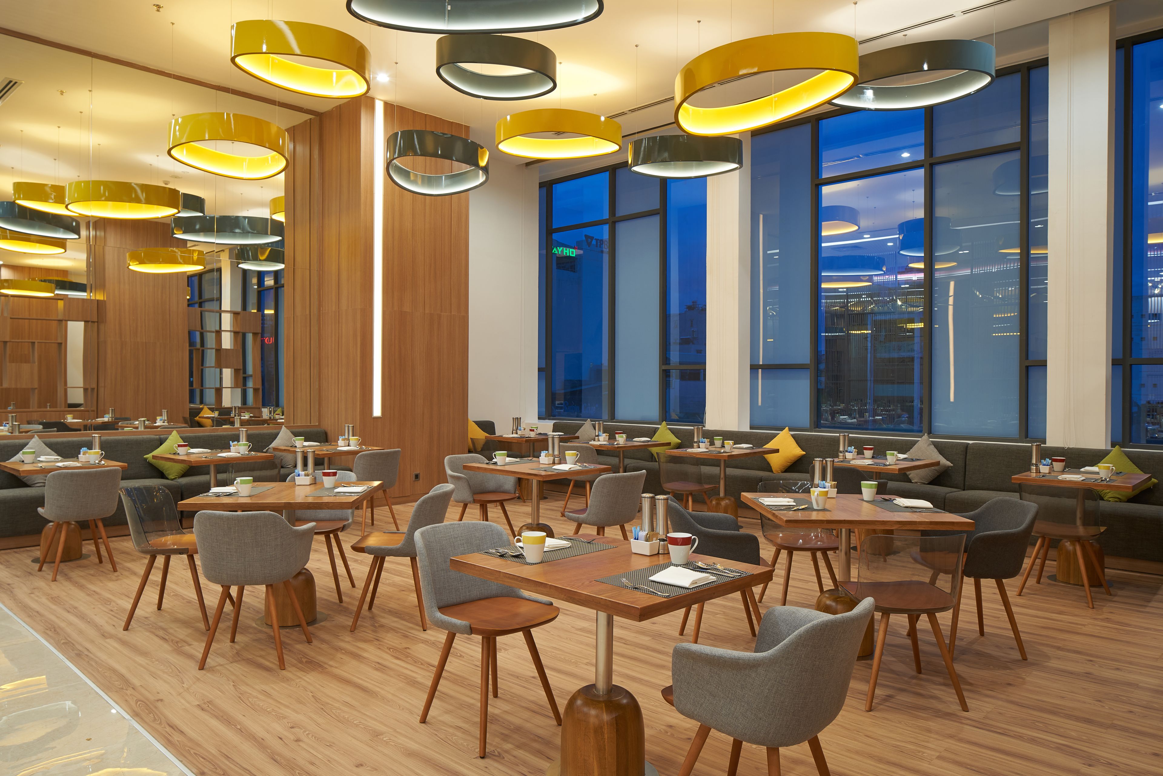 A modern dining area with multiple tables and chairs, large windows, and contemporary lighting fixtures.