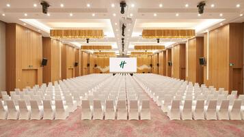 Un salón de conferencias con filas de sillas blancas, una pantalla grande al frente y paneles de madera en las paredes.
