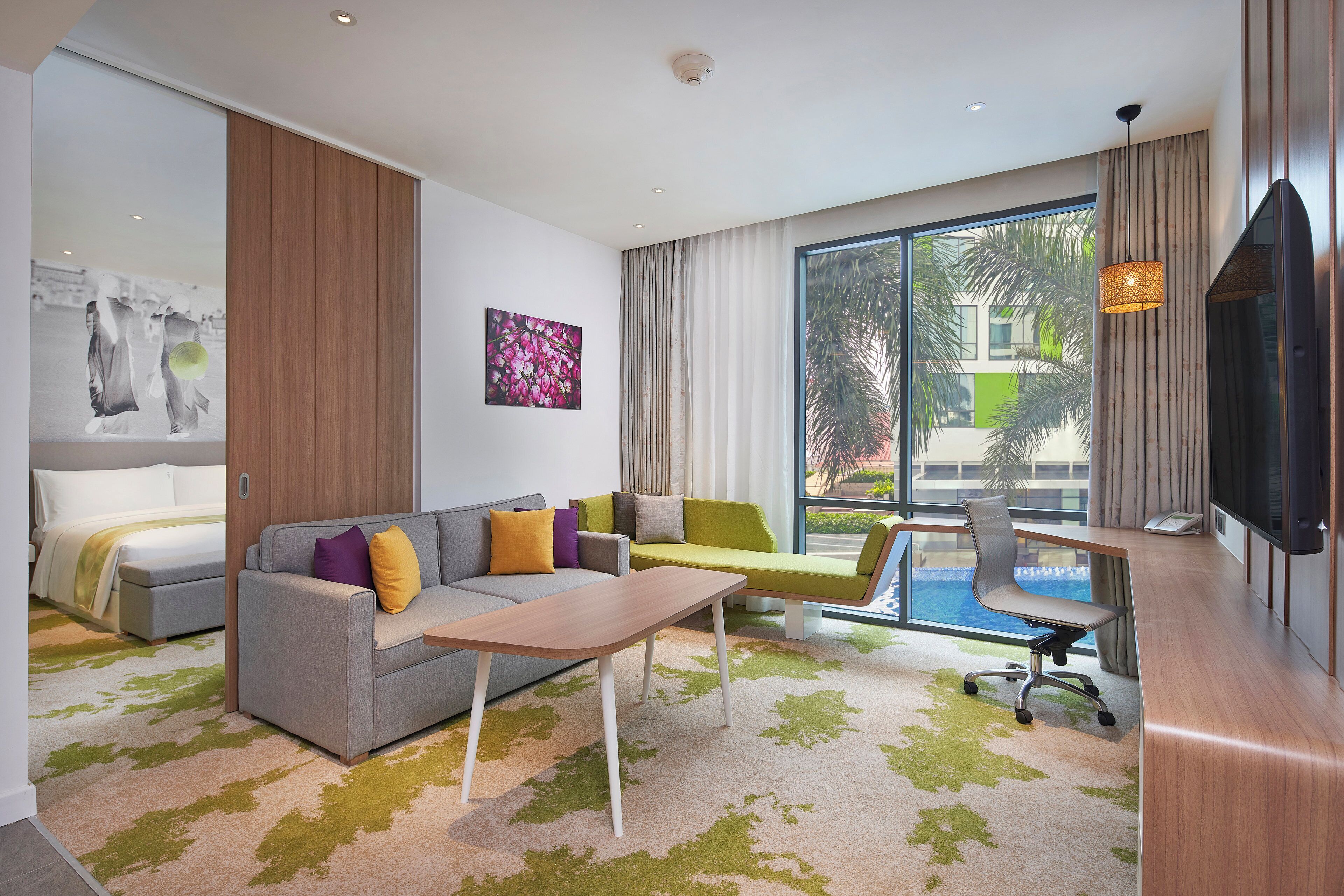 A modern hotel room with a grey sofa, a small table, a desk with a chair, and a large window with a view of a building and a pool.