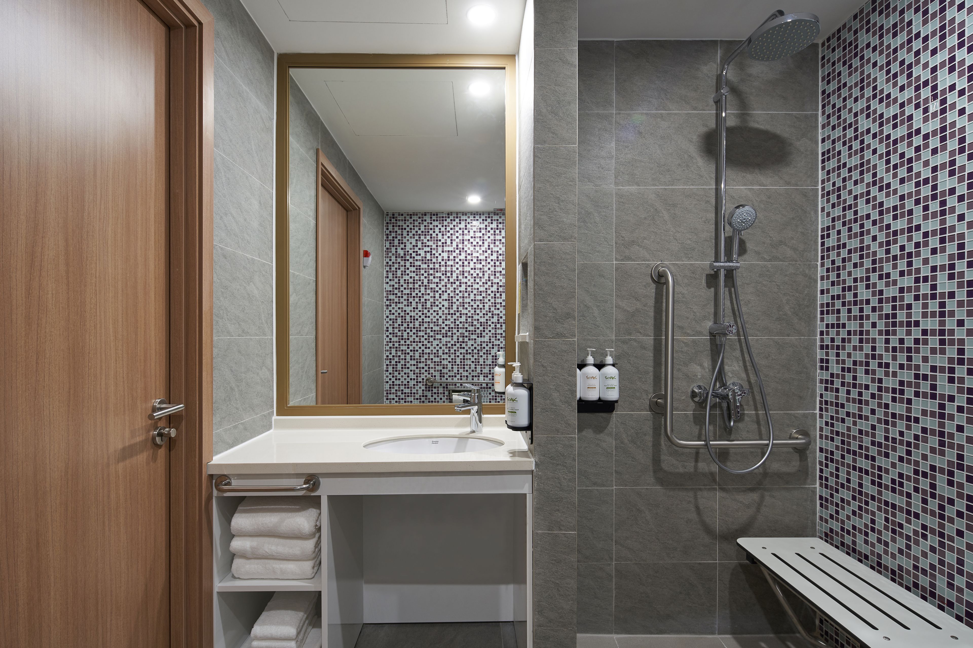 Ein modernes Badezimmer mit Holztür, Waschbecken in Weiß, Spiegel, Dusche mit Duschbrause und gefliesten Duschwänden.