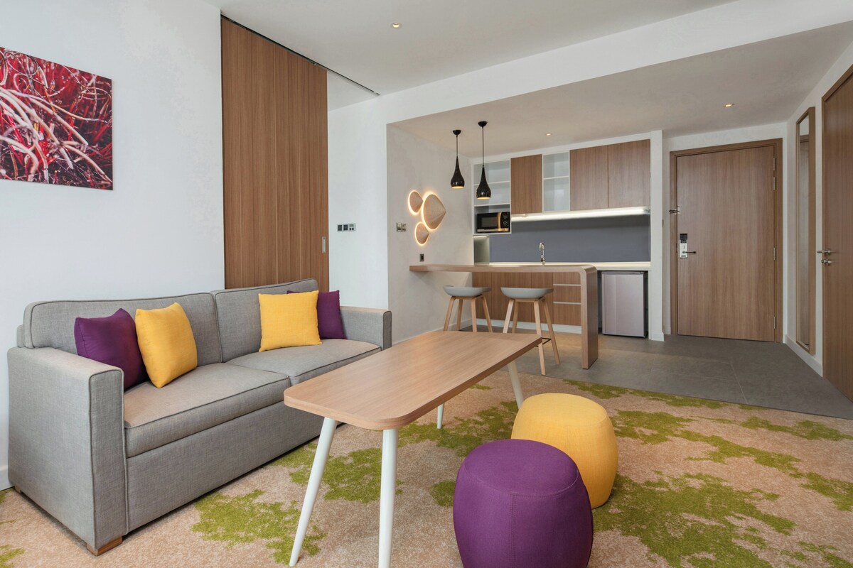 Een moderne woonkamer met een grijze bank, een houten salontafel en kleurrijke kussens.