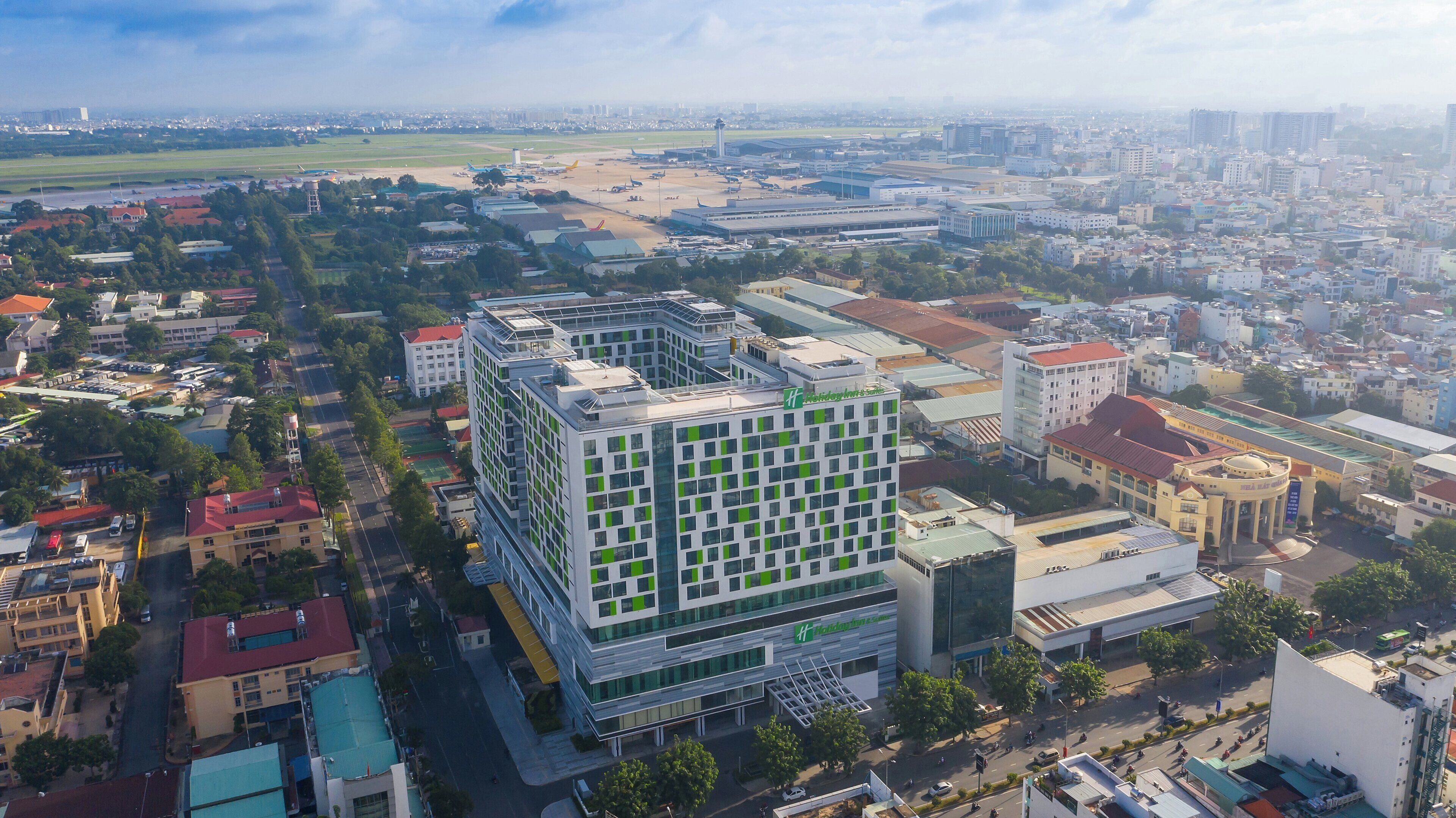 Un edificio moderno con un logotipo verde destacado, rodeado de edificios residenciales y comerciales, bajo un cielo despejado.