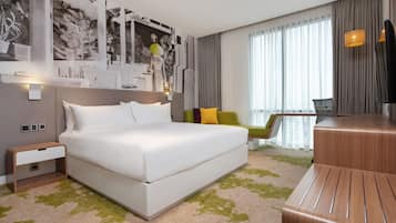 A modern hotel room with a large bed, a desk, a chair, and a large window with curtains.