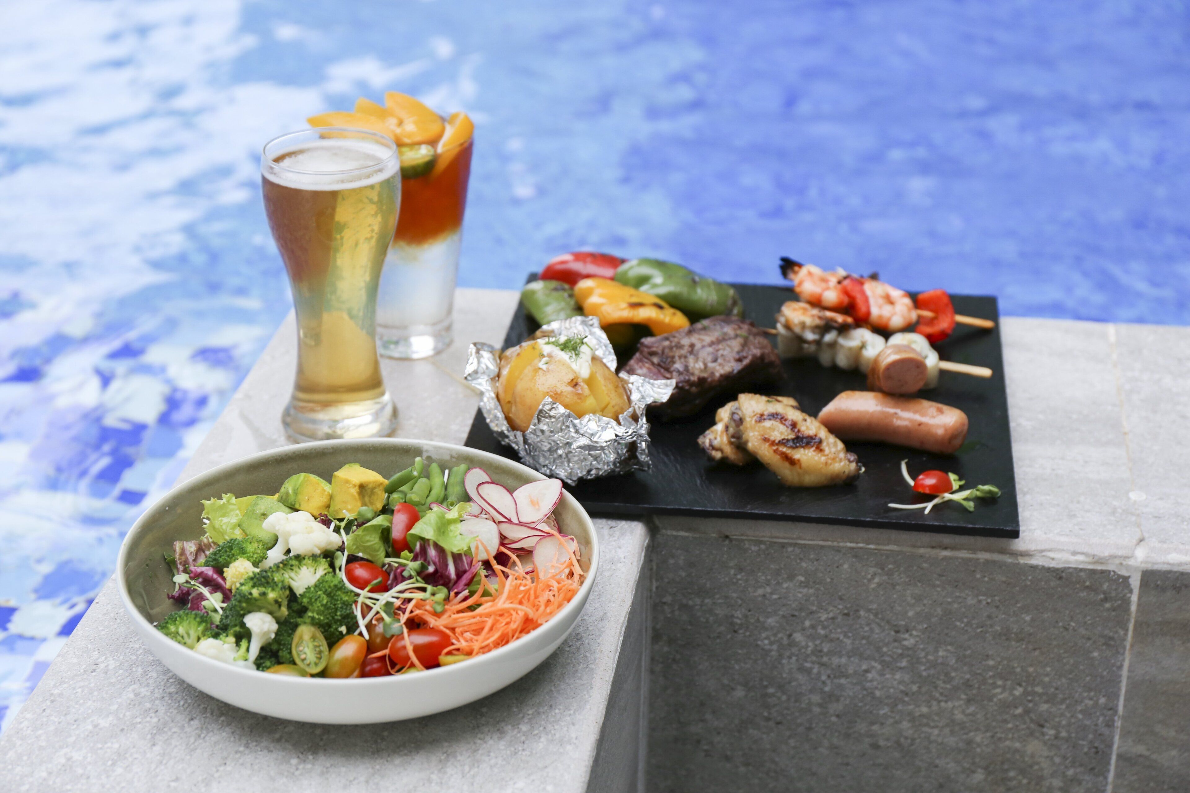 A dining table with a salad, grilled meats, and skewers, along with two glasses of beer.