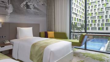 A modern hotel room with a large bed, a green armchair, a sofa, and a view of a swimming pool through a large window.