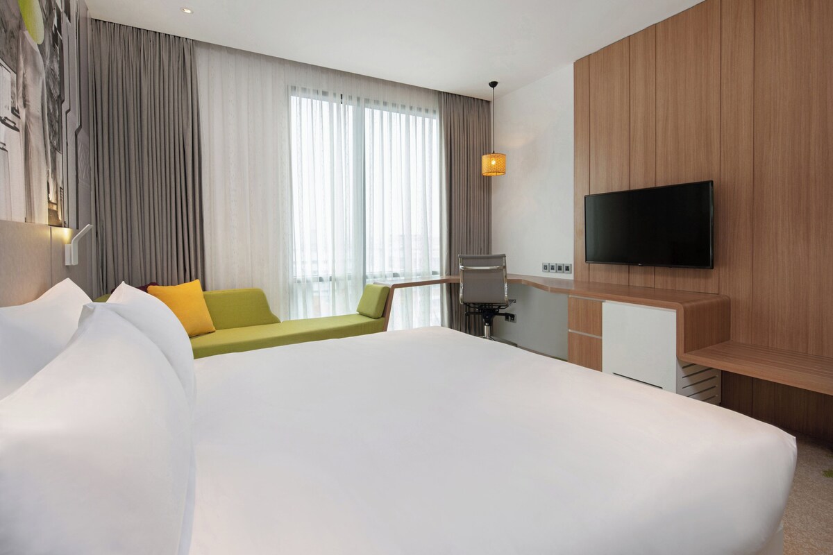 Een hotelkamer met een groot bed, een groene bank, een bureau met een flatscreen televisie en een raam met gordijnen.