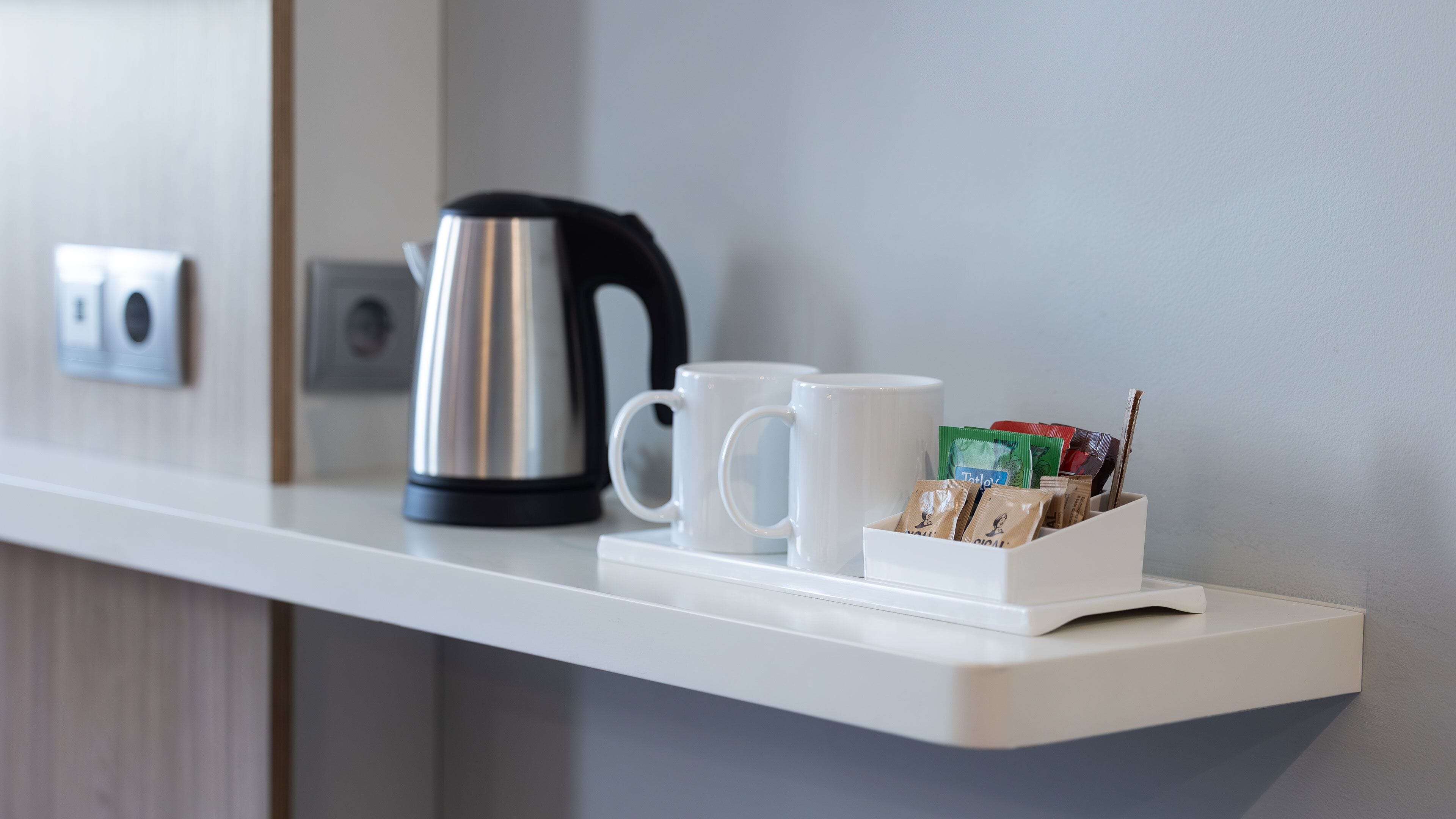 A white countertop with a kettle, two white mugs, and a dispenser with tea bags.