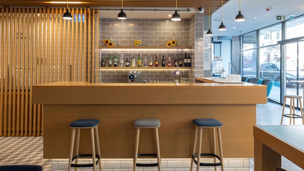 A bar area with a wooden counter, stools, and a variety of bottles and glasses. - Holiday Inn Express Porto City Center by IHG (Porto)