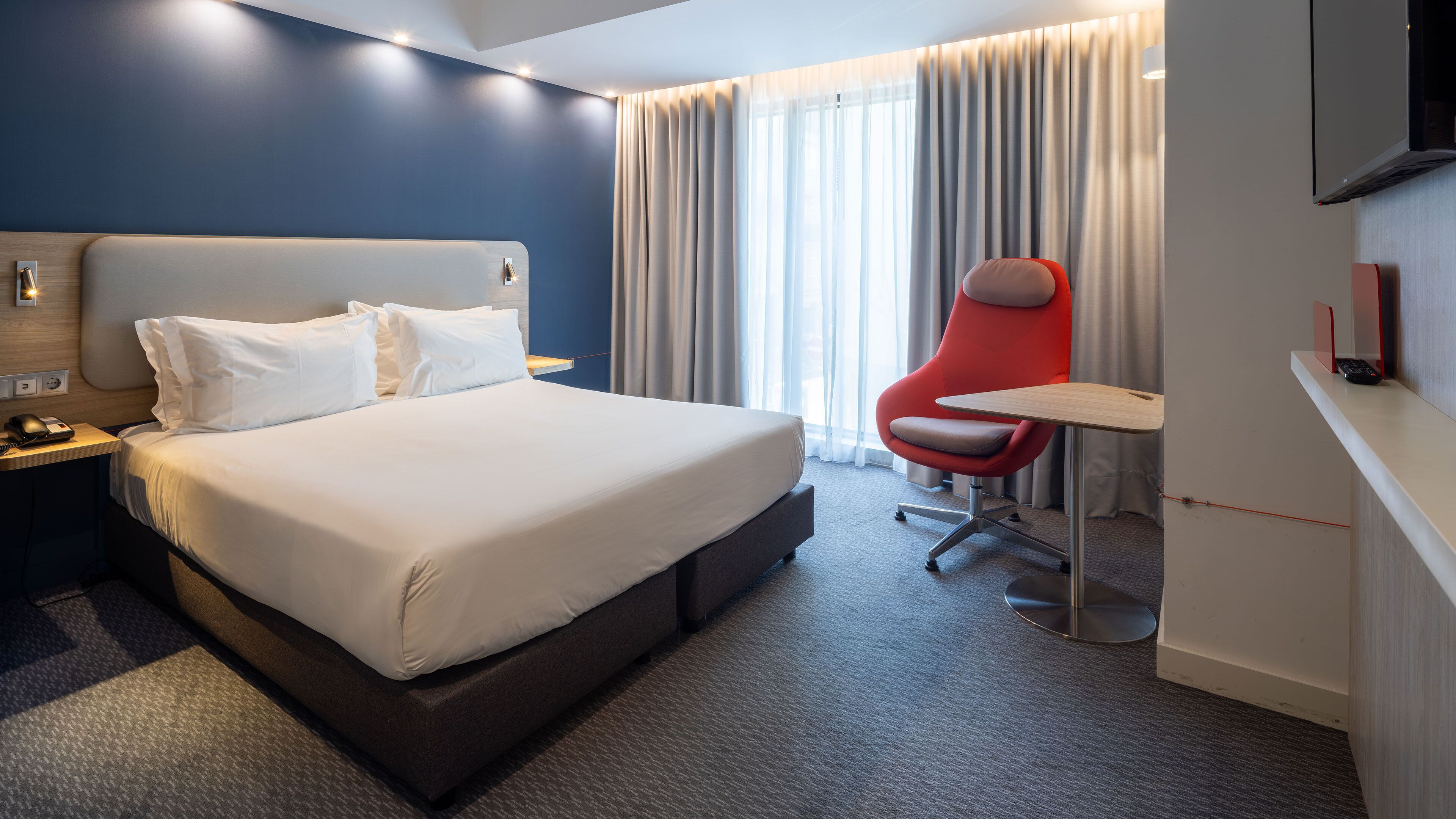 Una habitación de hotel con una cama grande, un escritorio, una silla roja y un televisor instalado en la pared.