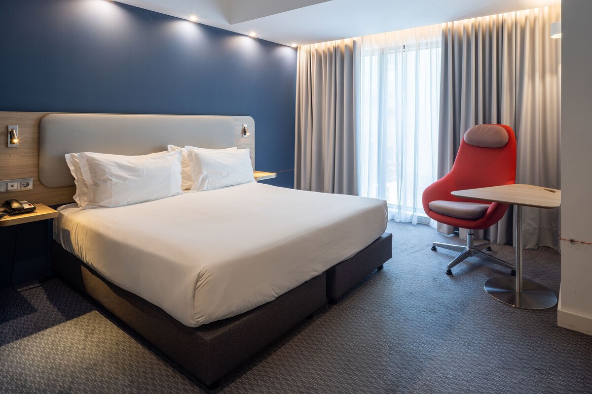 Une chambre d’hôtel avec un grand lit, un bureau, une chaise rouge et un téléviseur fixé au mur.