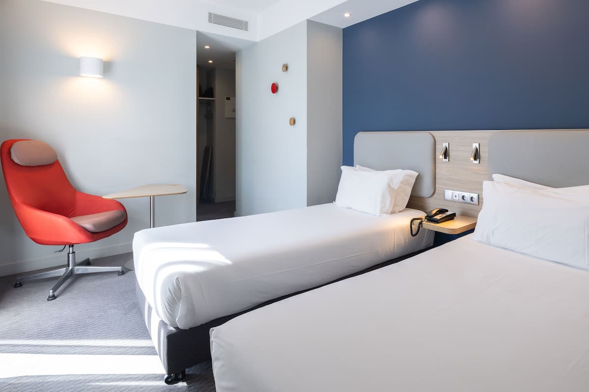 Hotellihuone, jossa on kaksi sänkyä, punainen tuoli, yöpöytä, jossa on puhelin, ja seinään kiinnitetty valaisin.